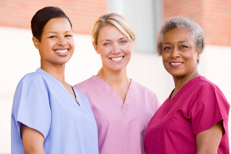 Three smiling nurses dressed in blue, pink, and red scrubs from left to right